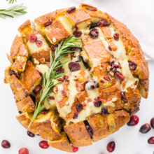Looking down on a loaf of bread cut into cubes with melted cheese, cranberries, and rosemary on top.