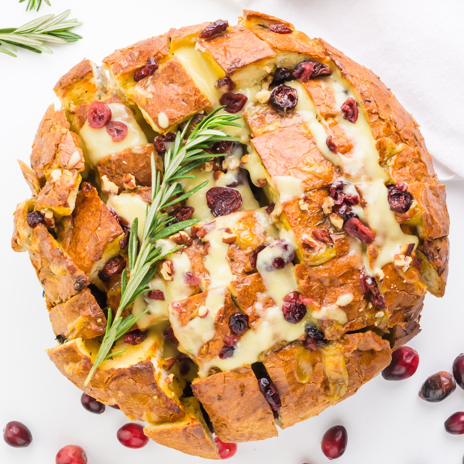 Looking down on a loaf of bread cut into cubes with melted cheese, cranberries, and rosemary on top.