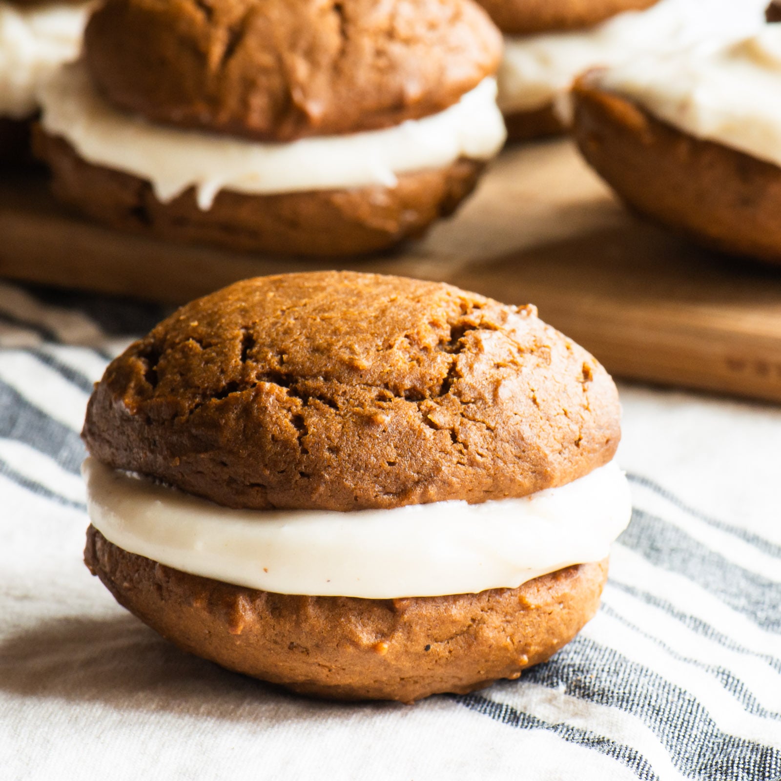 A gingerbread whoopie pie sits in front of a wooden board with more cookies on it.