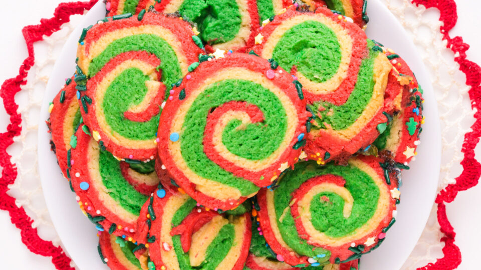 A plate of Christmas pinwheel cookies sits on a white plate and a red and white doily.