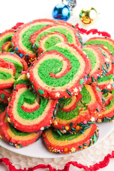 A plate of Christmas color inspired pinwheel cookies sits next to colorful christmas ornaments.