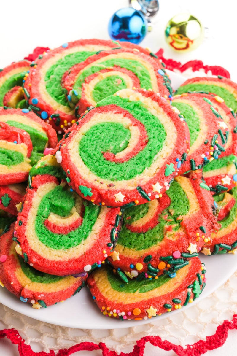 A plate of Christmas color inspired pinwheel cookies sits next to colorful christmas ornaments.