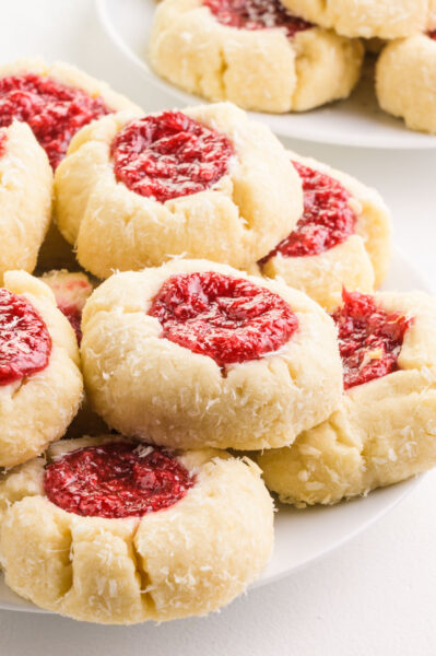 Raspberry thumbprint cookies are stacked on a plate in front of another plate with more of the cookies.