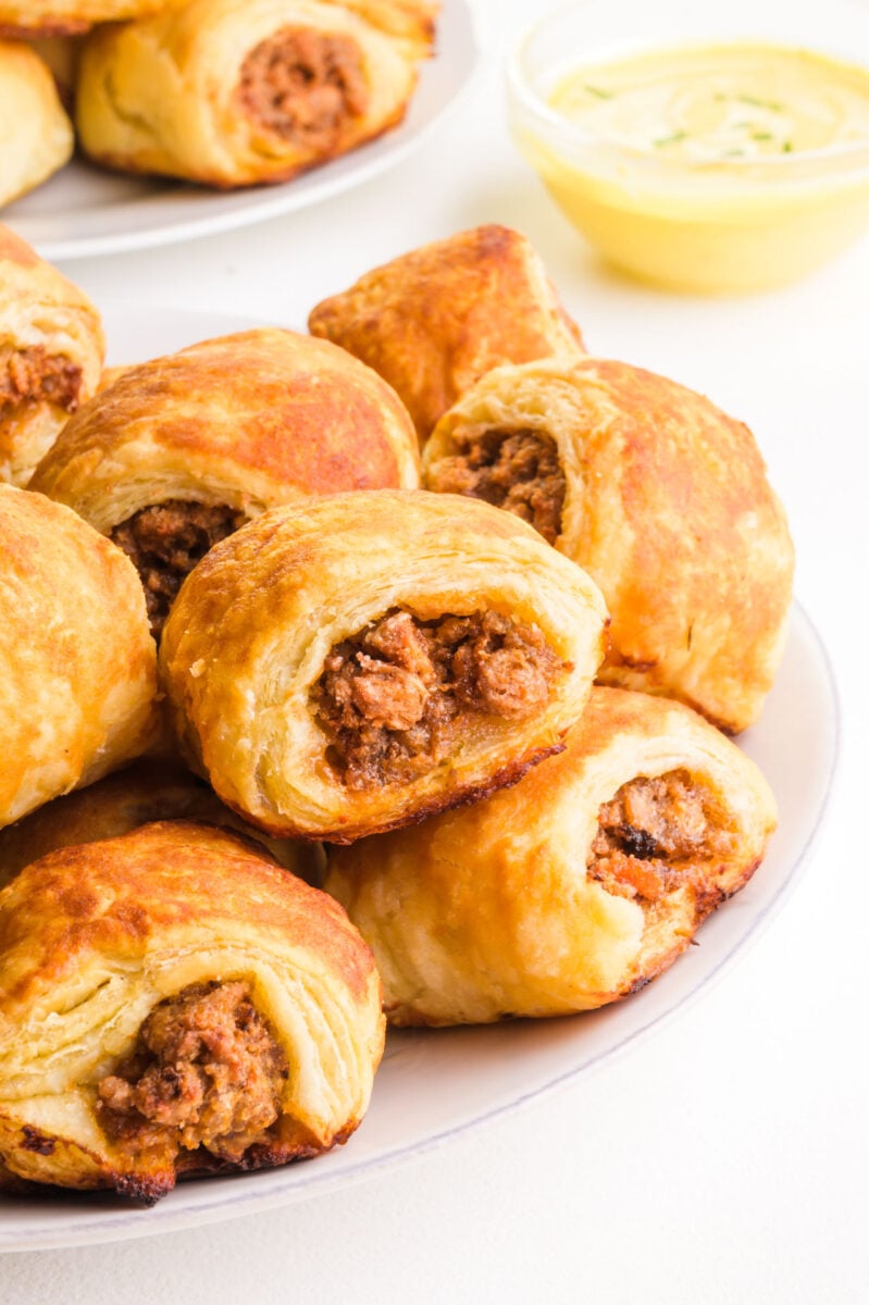 A plate of vegan sausage rolls sits in front of another plate of the treats in the background and a bowl of mustard sauce.