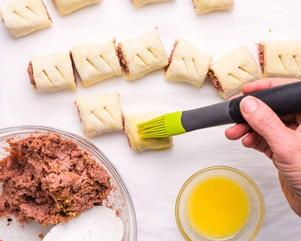 A hand brushes vegan egg wash over vegan sausage rolls next to a bowl with more vegan sauce mixture.