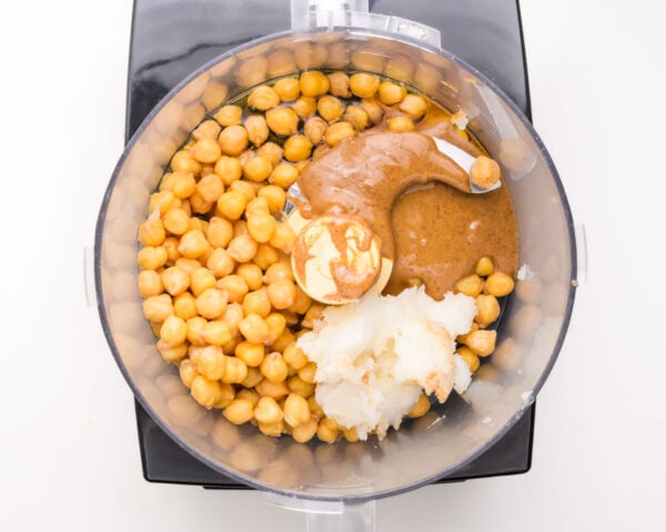 A food processor is full of ingredients like chickpeas, maple syrup, and coconut oil.