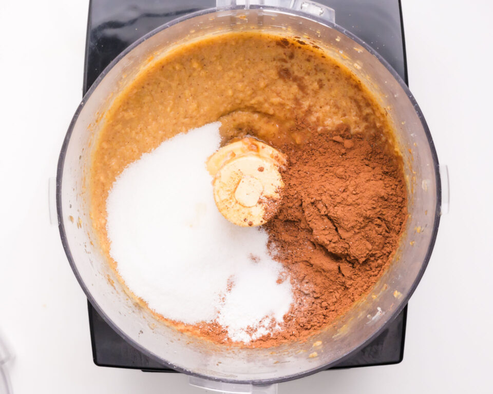 A food processor is full of ingredients like a mixed chickpea batter, cocoa powder, and sugar.