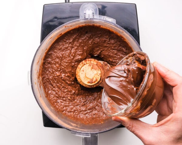 Melted chocolate is being poured into a food processor with chocolate brownie batter in it.