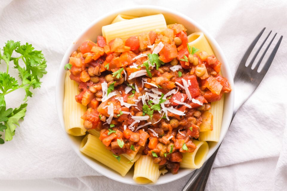 A bowl of pasta is topped with lentil bolognese and sits next to a fork.