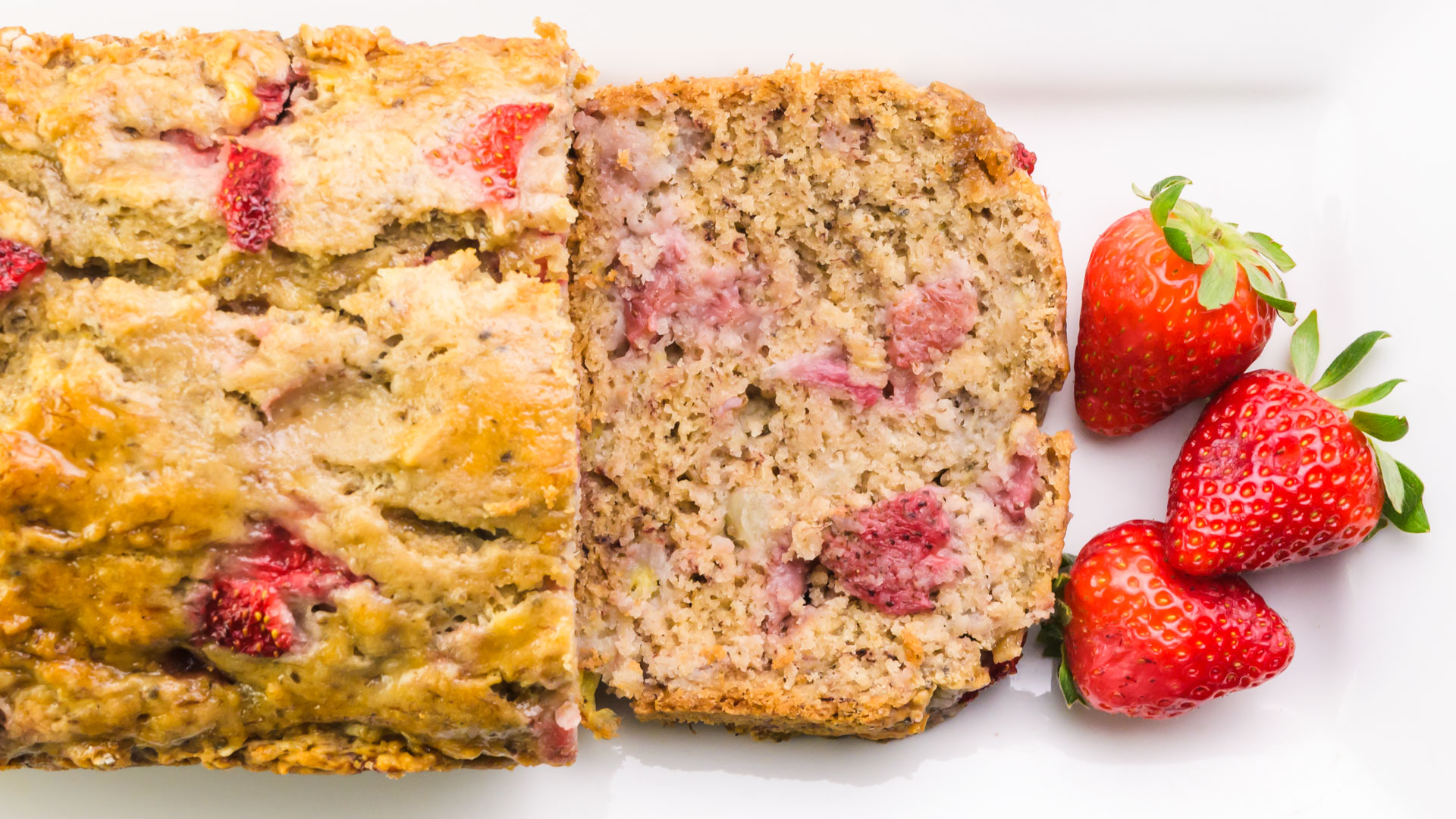 Looking down on a loaf of strawberry banana bread with one slice laying in front of it along with fresh strawberries.