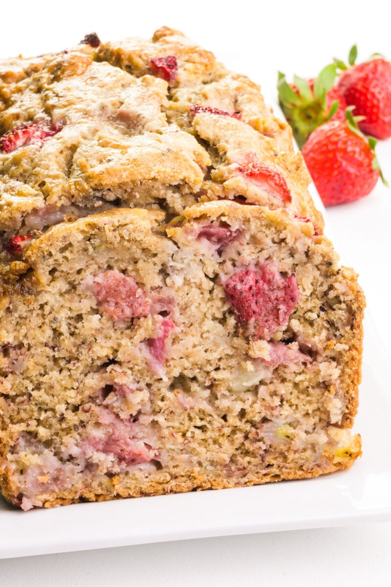 A loaf of vegan strawberry banana bread on a plate sits next to fresh strawberries.