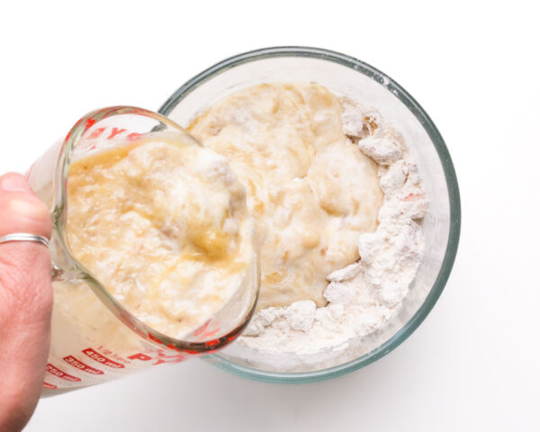 A mashed banana mixture is being poured into a bowl with a flour mixture.