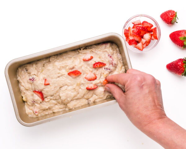 A hand places chopped strawberries on top of batter in a loaf pan.