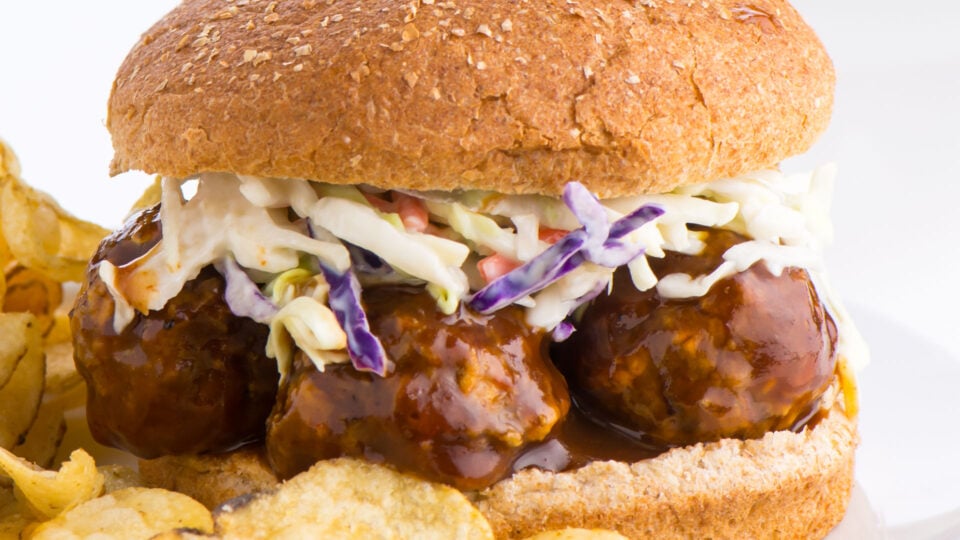 A BBQ meatball sandwich has coleslow and sits next to potato chips on a plate.