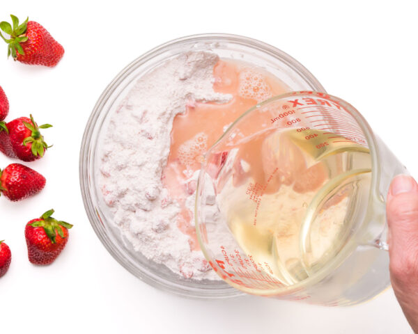 Wet ingredients are being poured into a mixing bowl. Fresh strawberries are by the bowl.