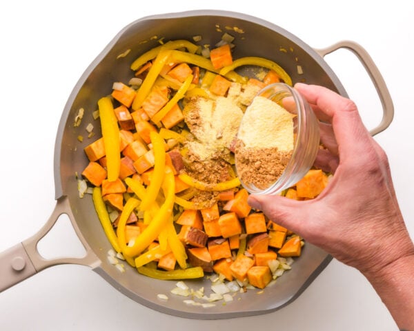 Spices are being poured into a skillet with onions, peppers, and more.