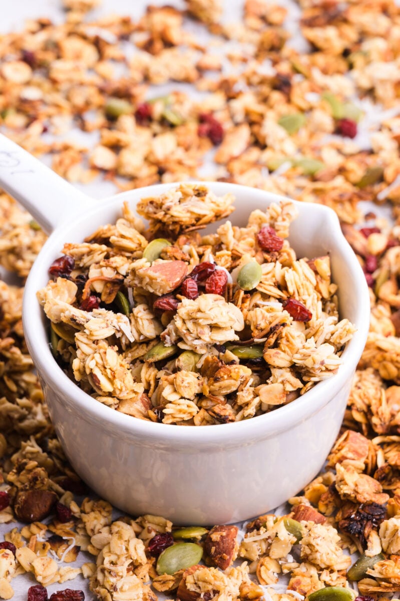 A measuring cup holds granola in a pan with more granola.