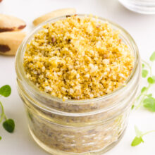 A jar filled with finely ground yellow and white nut mixture, surrounded by pine nuts and green herbs.