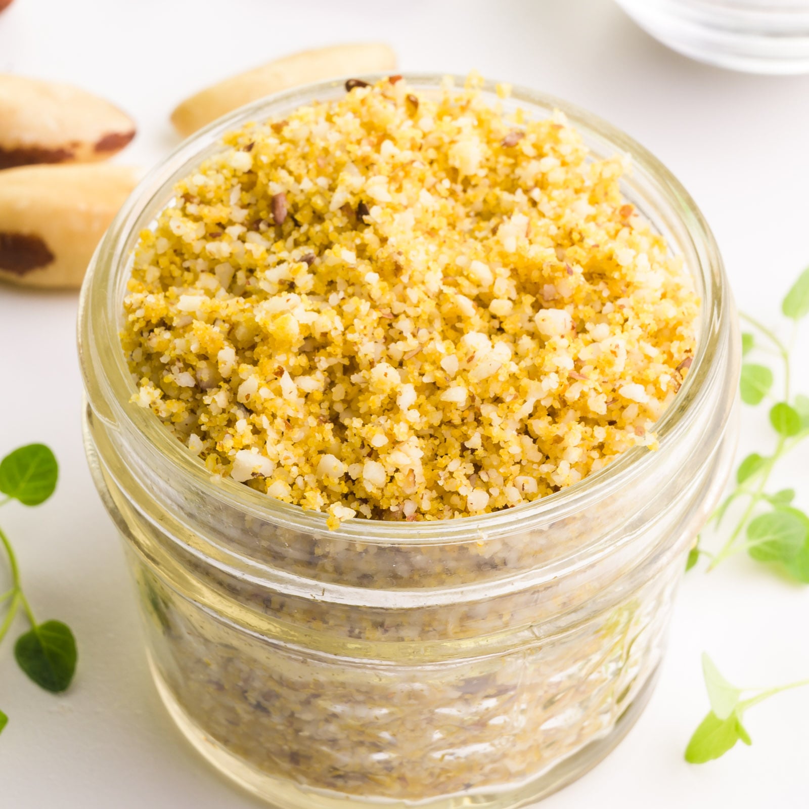 A jar filled with finely ground yellow and white nut mixture, surrounded by pine nuts and green herbs.