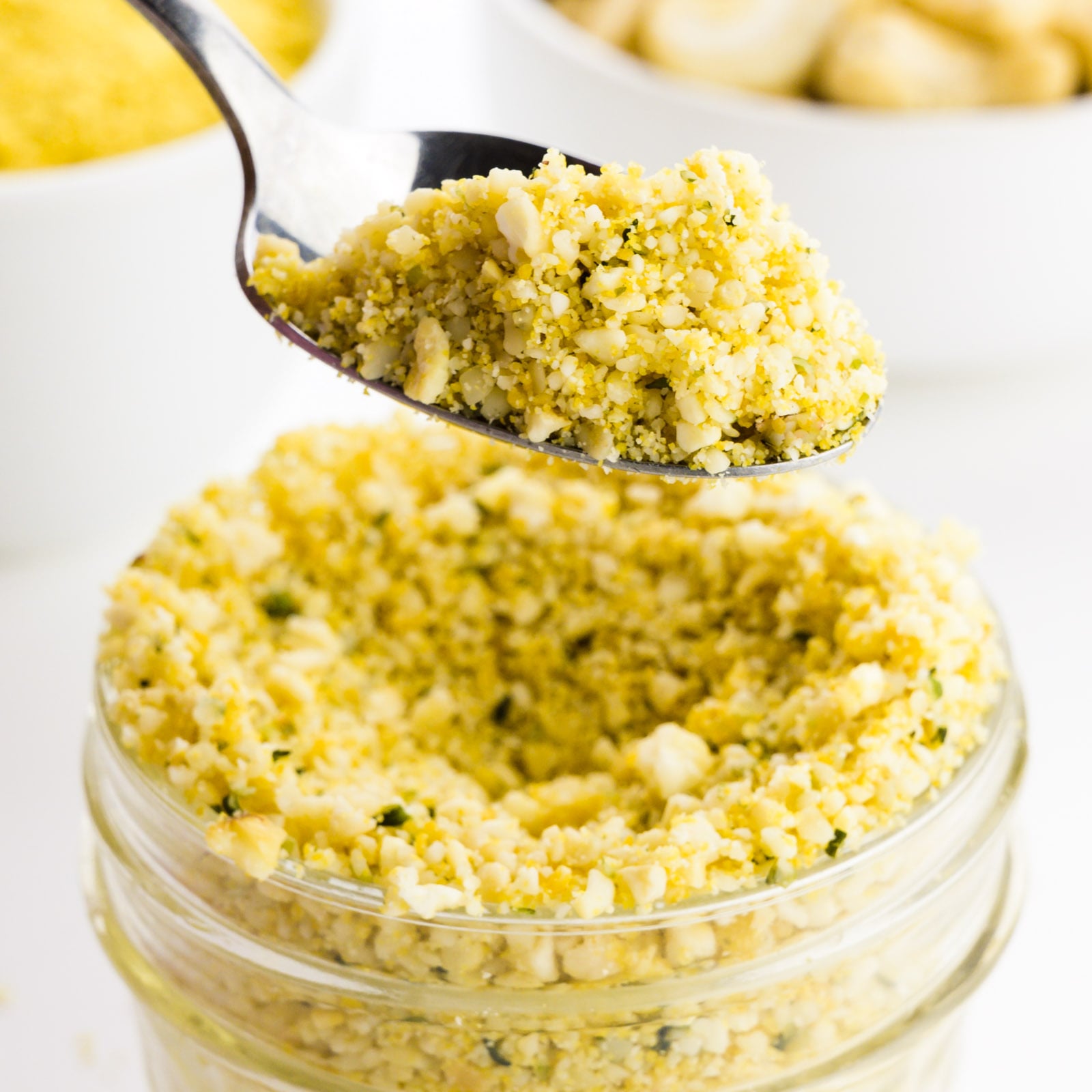 A hand holds a spoonful of cashew parmesan having over a glass jar full of it wit cashews behind it.