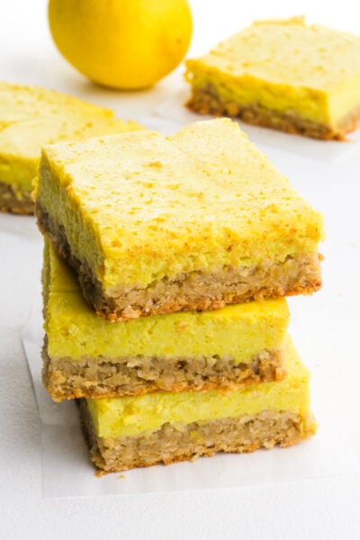 A stack of lemon bars on a white table sits near more of the dessert slices and a lemon.
