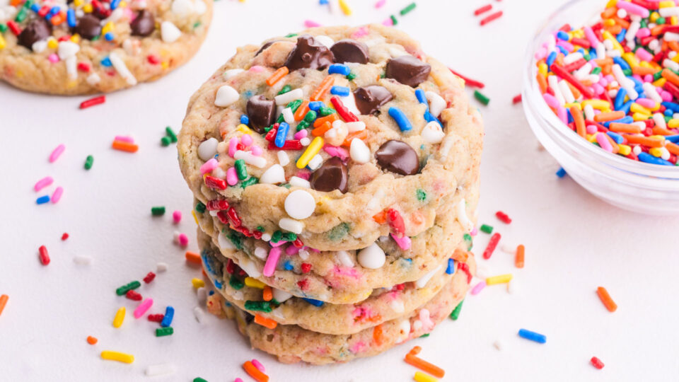 A stack of chocolate chip sprinkle cookies have sprinkles all around.