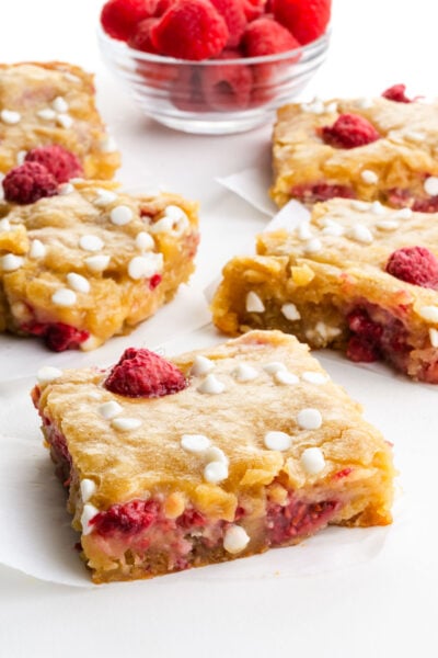 Slices of raspberry white chocolate dessert bars on a table next to a bowl of fresh raspberries.