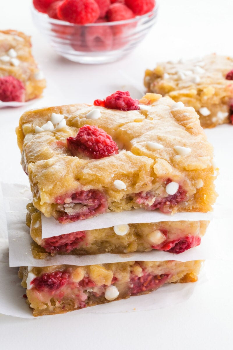 A stack of raspberry blondies sit in front of more dessert slices and fresh raspberries.