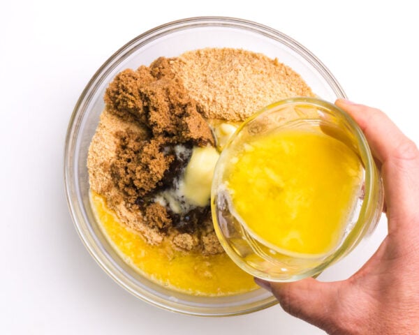 Melted butter is being poured into a bowl with graham cracker crumbs and other ingredients.