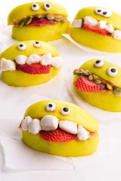 Several apple monster treats are on a table, with candy eyes and marshmallows for teeth.