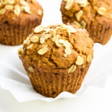 Three apple muffins sit on a white counter. They all show bits of oats on top. The front one is in focus.