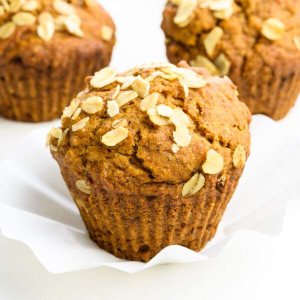 Three apple muffins sit on a white counter. They all show bits of oats on top. The front one is in focus.