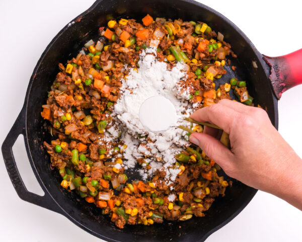 Flour is being sprinkled over veggies in an iron skillet.