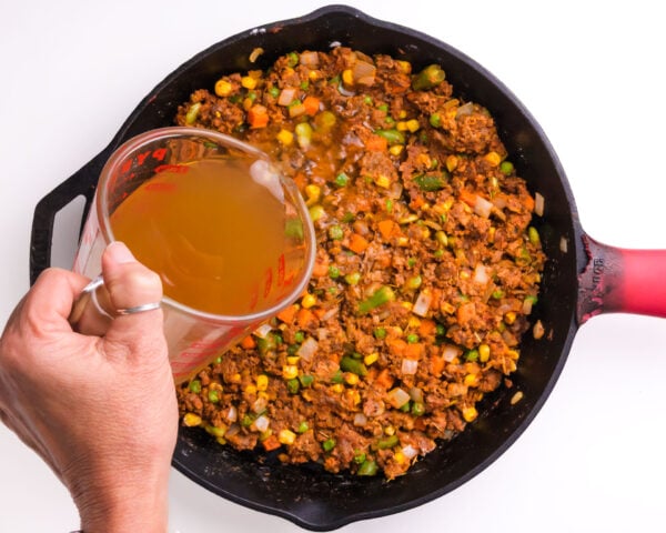 Broth is being poured into a skillet with vegetables and other ingredients.