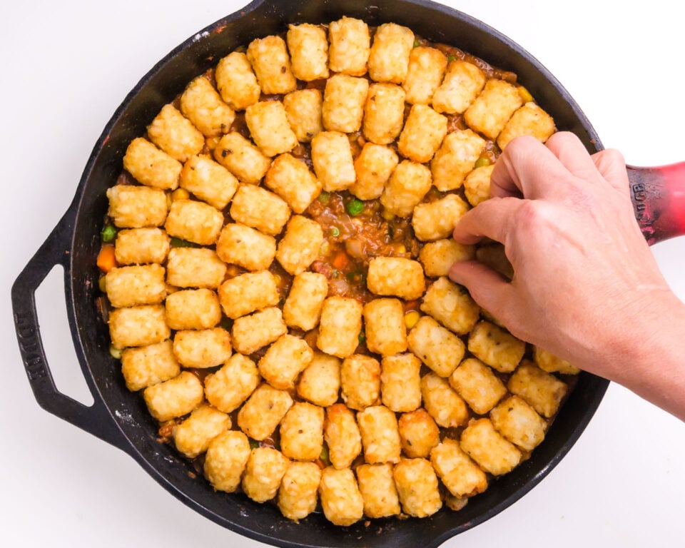 Tater tots are being arranged on top of a vegetable mixture in a skillet.
