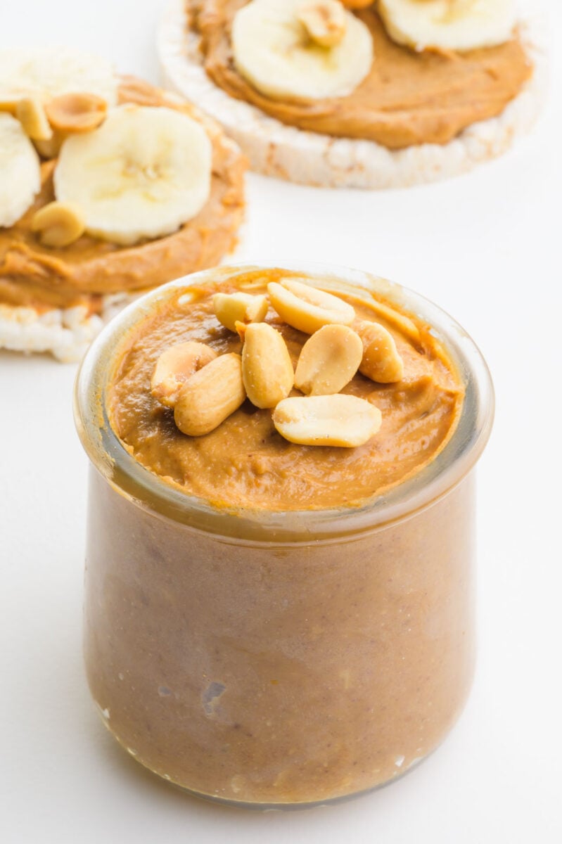 A jar of peanut butter has nuts on top sitting in front of rice cake snacks.