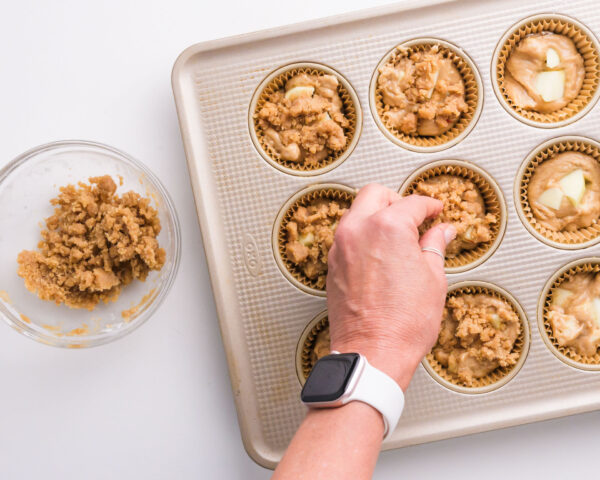Crumble topping is being added to batter in muffin compartments.