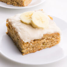 Slice of banana cake topped with cream cheese frosting and banana slices, served on a white plate.