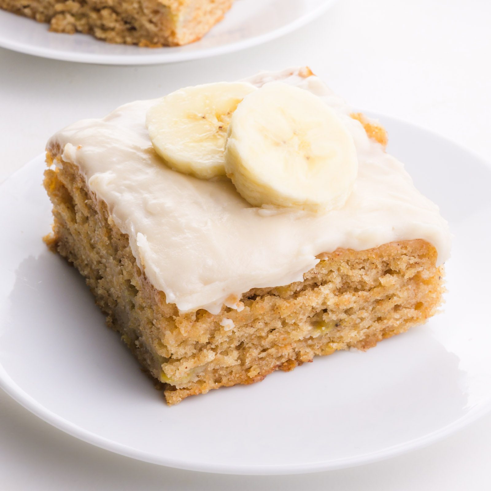 Slice of banana cake topped with cream cheese frosting and banana slices, served on a white plate.