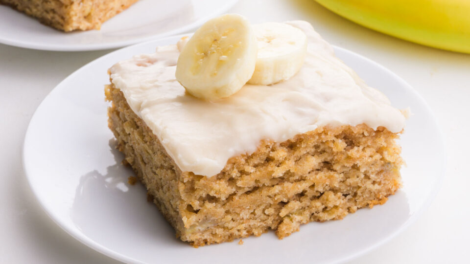 Banana snack cake on a plate topped with frosting and banana slices.