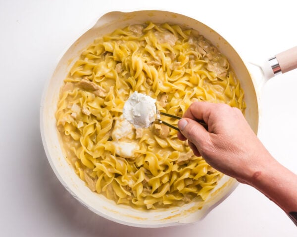 Vegan cream cheese i being spooned into a skillet with noodles and other ingredients.