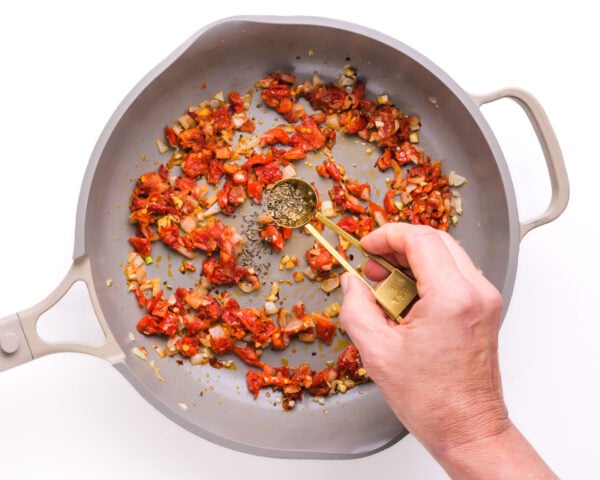Spices are being added to a skillet with other ingredients, like sun-dried tomatoes.