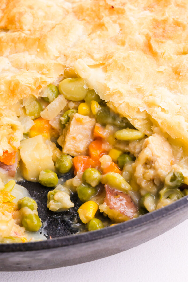 A pot pie in a skillet has a golden crust, with a bite taken out revealing creamy veggies inside.
