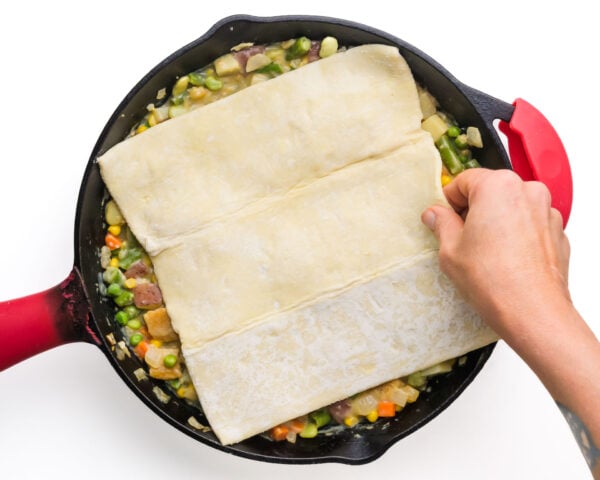 A hand placing dough over mixed vegetables in a skillet with a red handle.