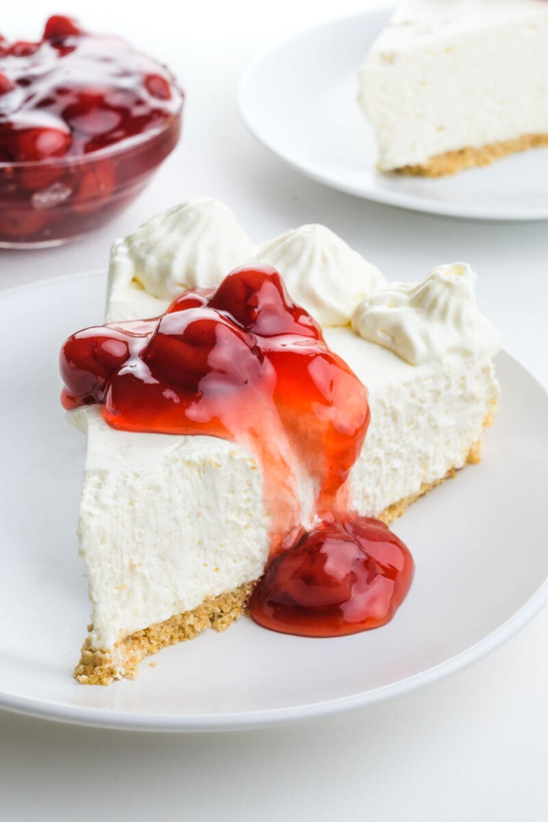 A slice of cheesecake on a plate is tilted to the side showing whipped cream and cherry sauce on top.