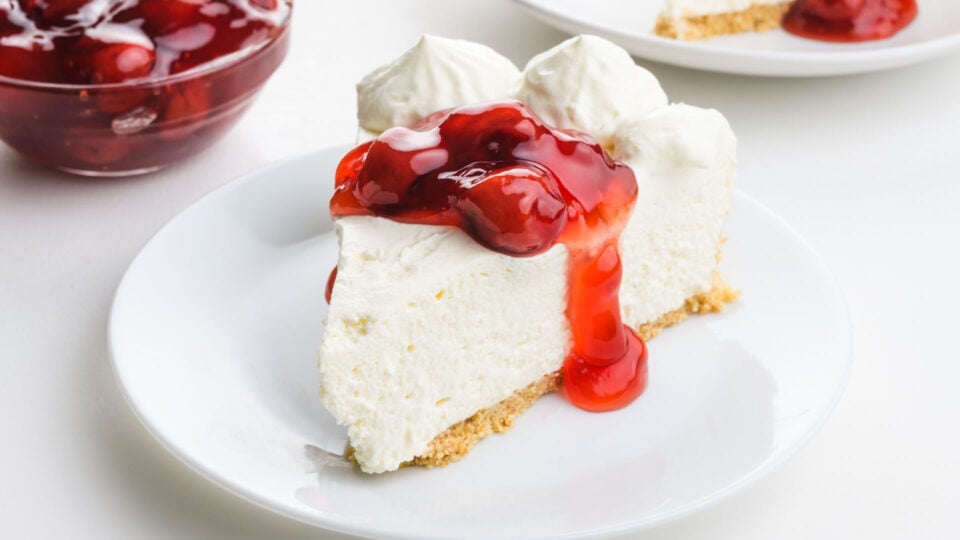 A slice of cheesecake on a plate is topped with whipped cream and cherry sauce.