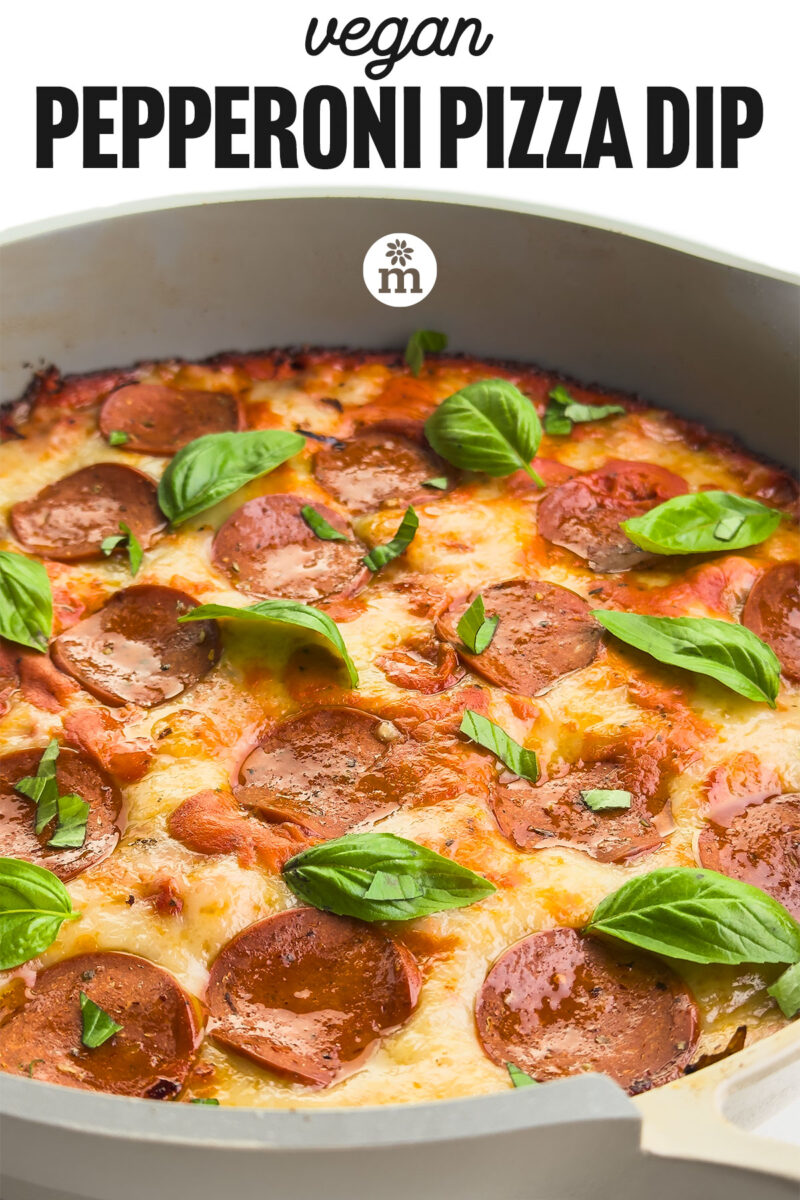 Vegan pepperoni pizza dip with melted cheese, topped with fresh basil leaves in a baking dish.