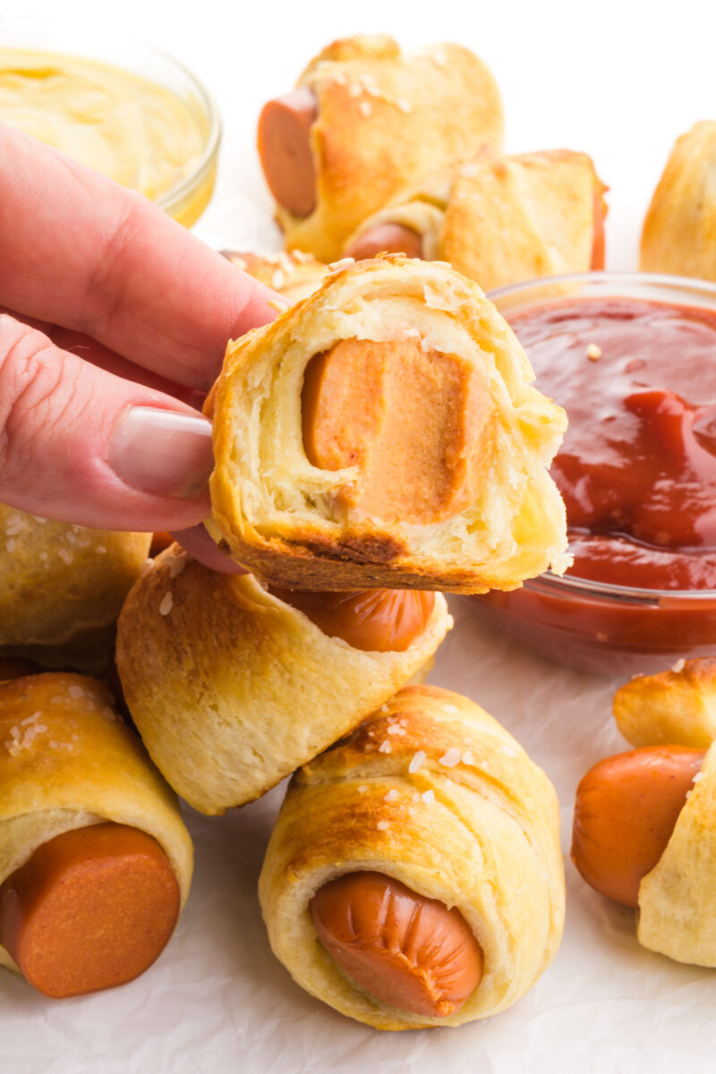 A hand holds a pig in a blanket with a bite taken out, revealing a hot dog inside.