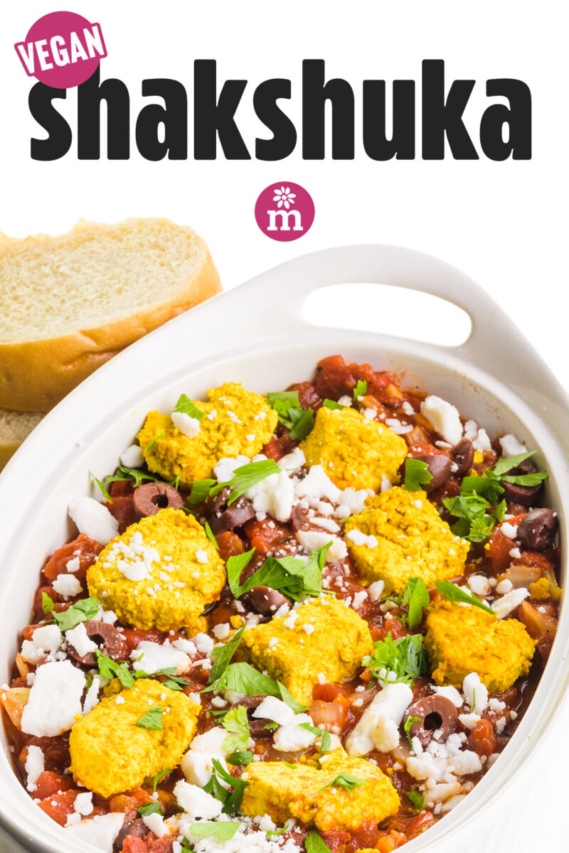 Vegan shakshuka with golden topping, herbs, olives, and white cheese, served alongside slices of bread.