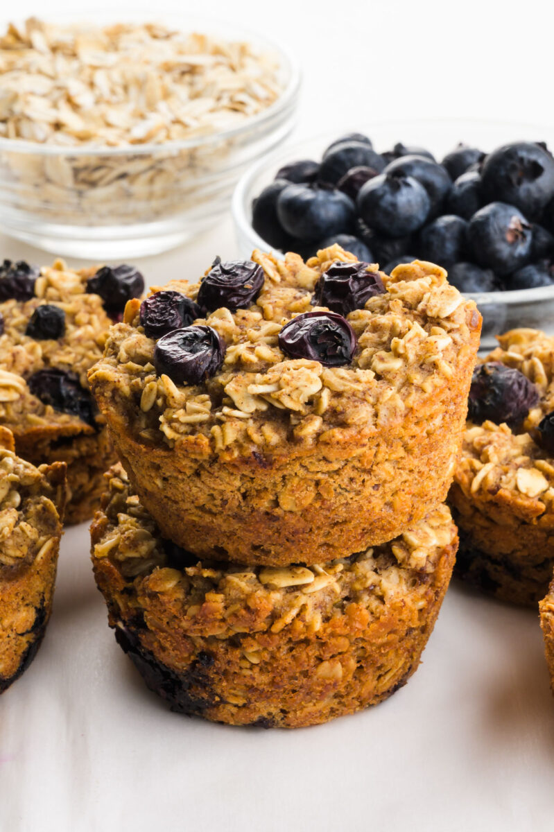 A stack of blueberry oatmeal cups sits in front of fresh blueberries and a bowl of oats.